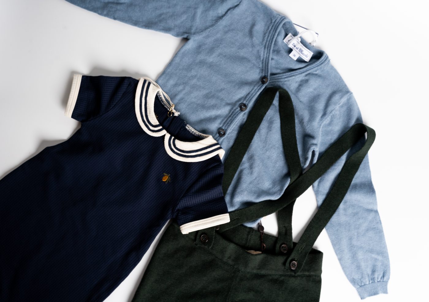 Flat-lay of organic baby clothes in the Filiz box — navy dress, blue cardigan, green corduroy overalls